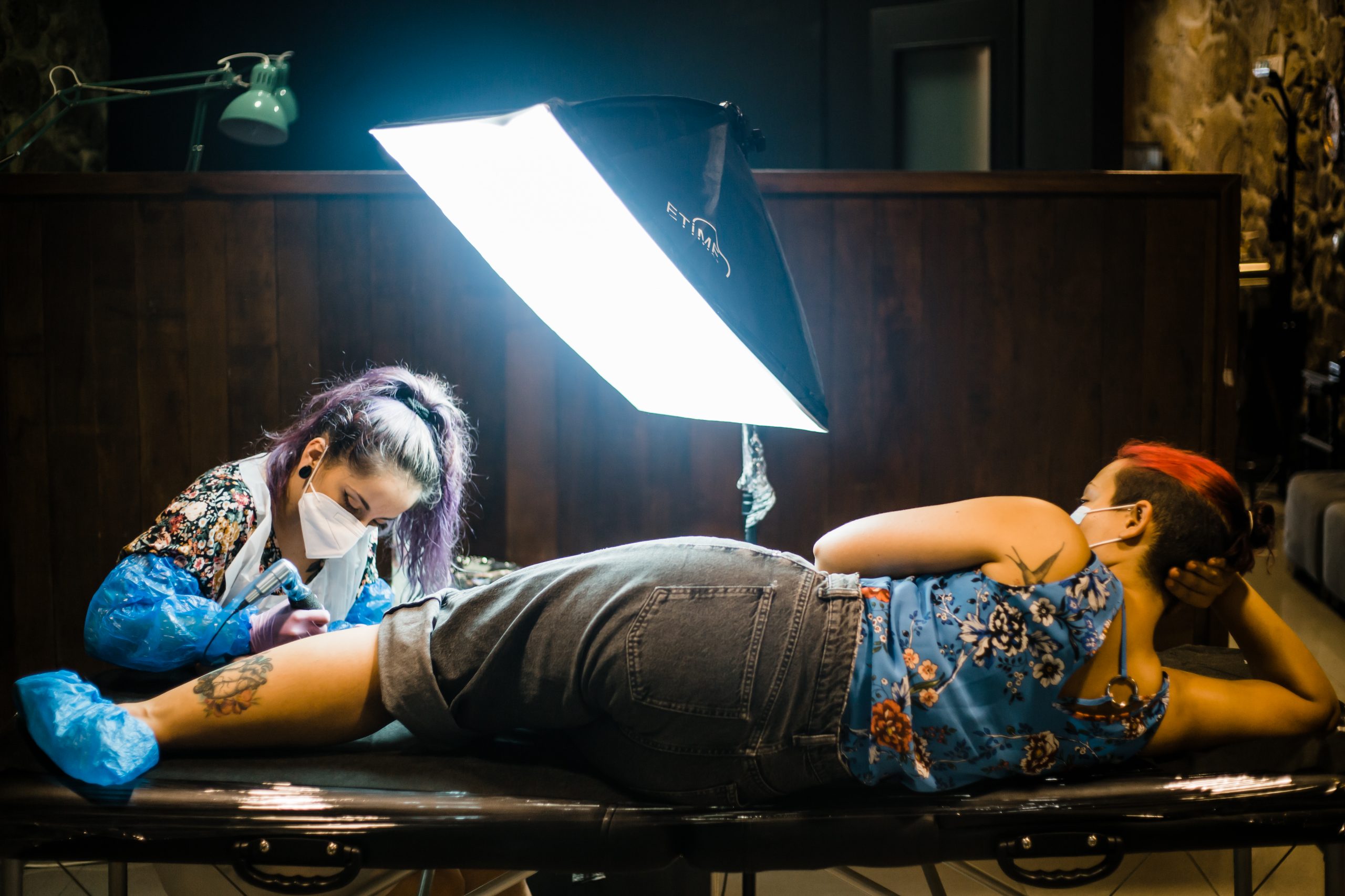 mulher sentada em marquesa a ser tatuada na perna por uma tatuadora